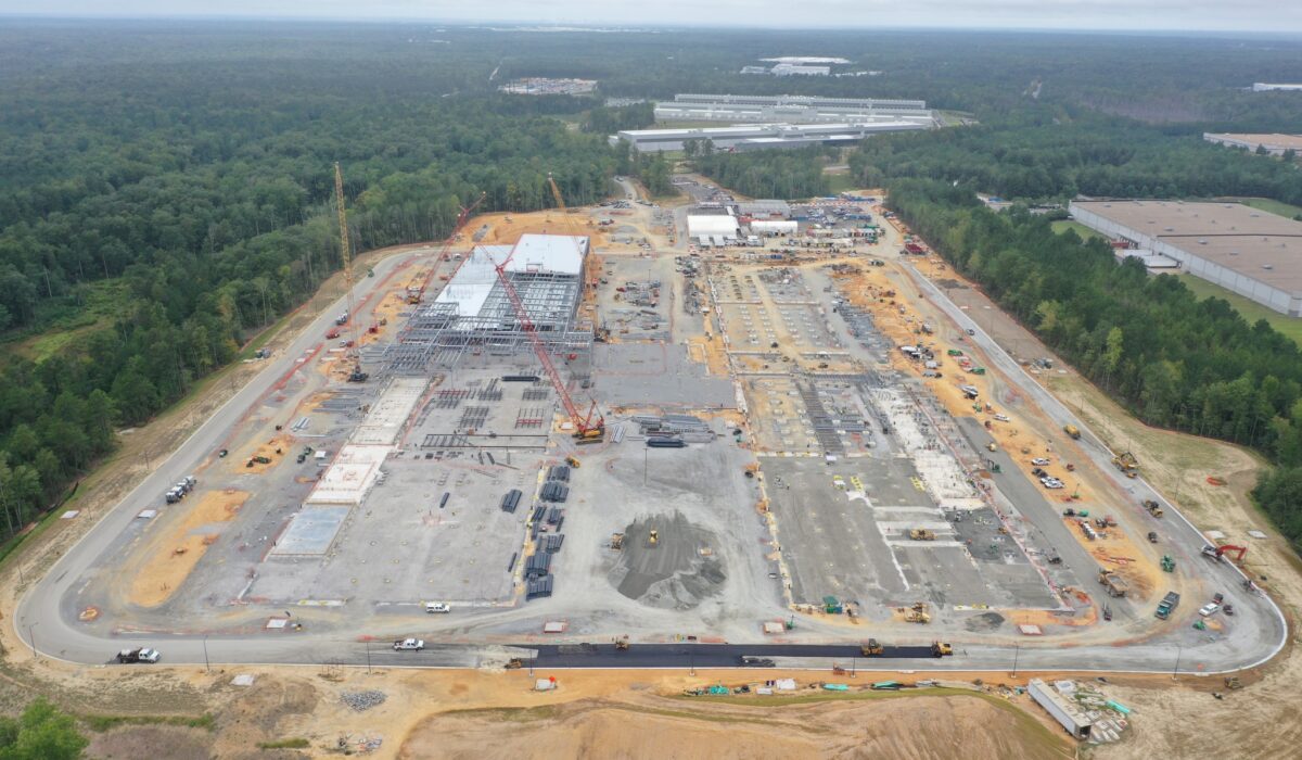 Aerial view: construction site, steel frames, cranes, forest nearby.