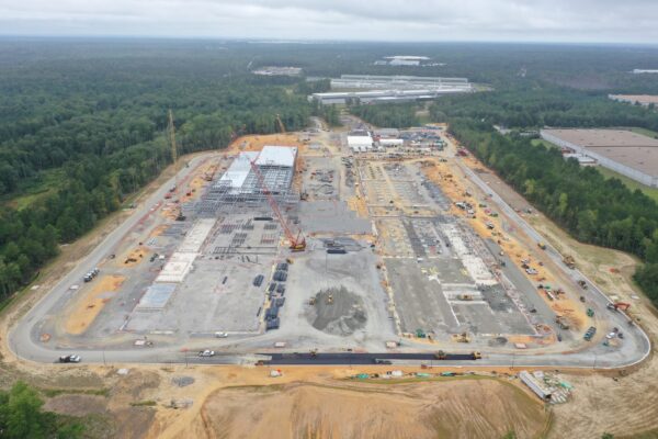 Aerial view: construction site, steel frames, cranes, forest nearby.