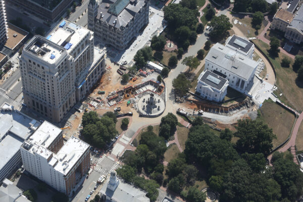 Aerial view: construction site, central monument, streets, and trees.