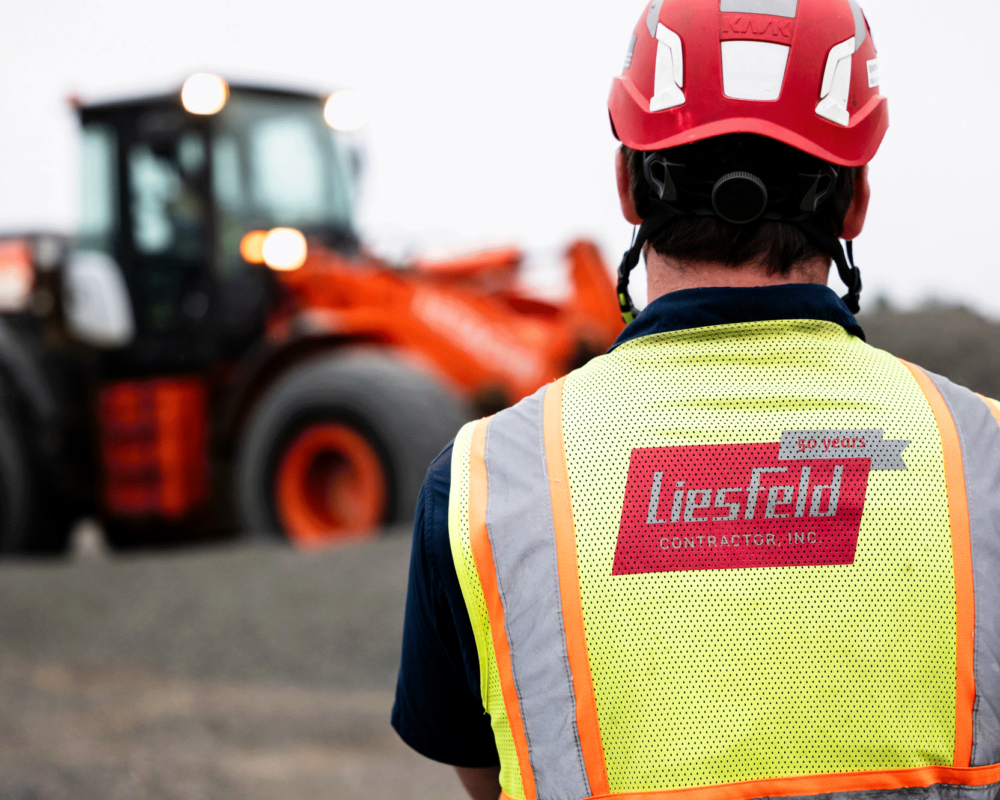 Worker in red hard hat faces blurred orange construction vehicle.