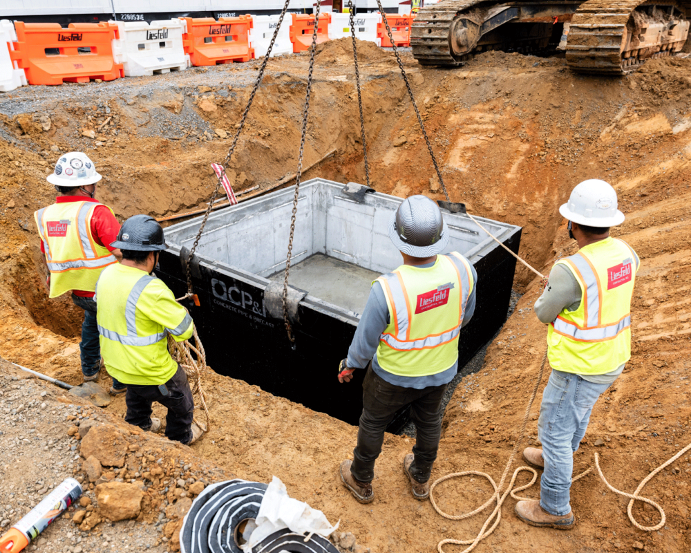 Workers guide precast concrete structure into hole, crane assists, barriers surround.