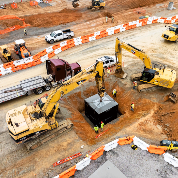 Aerial view: two excavators lift container, workers and equipment nearby.