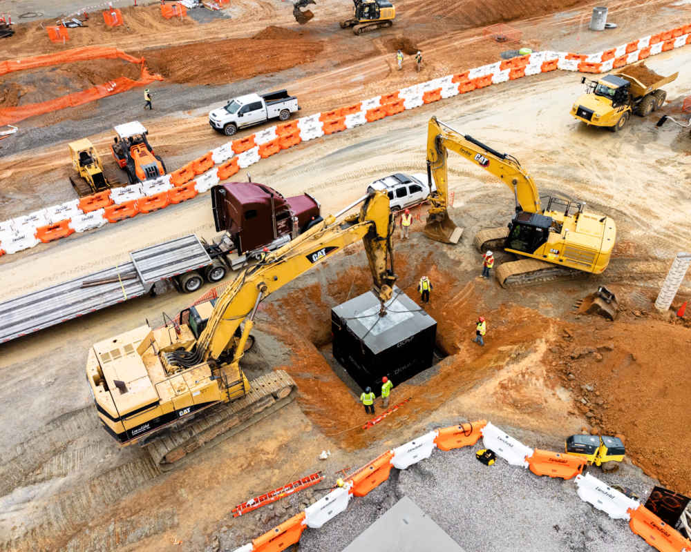 Aerial shot: two excavators lower black precast concrete structure into pit.