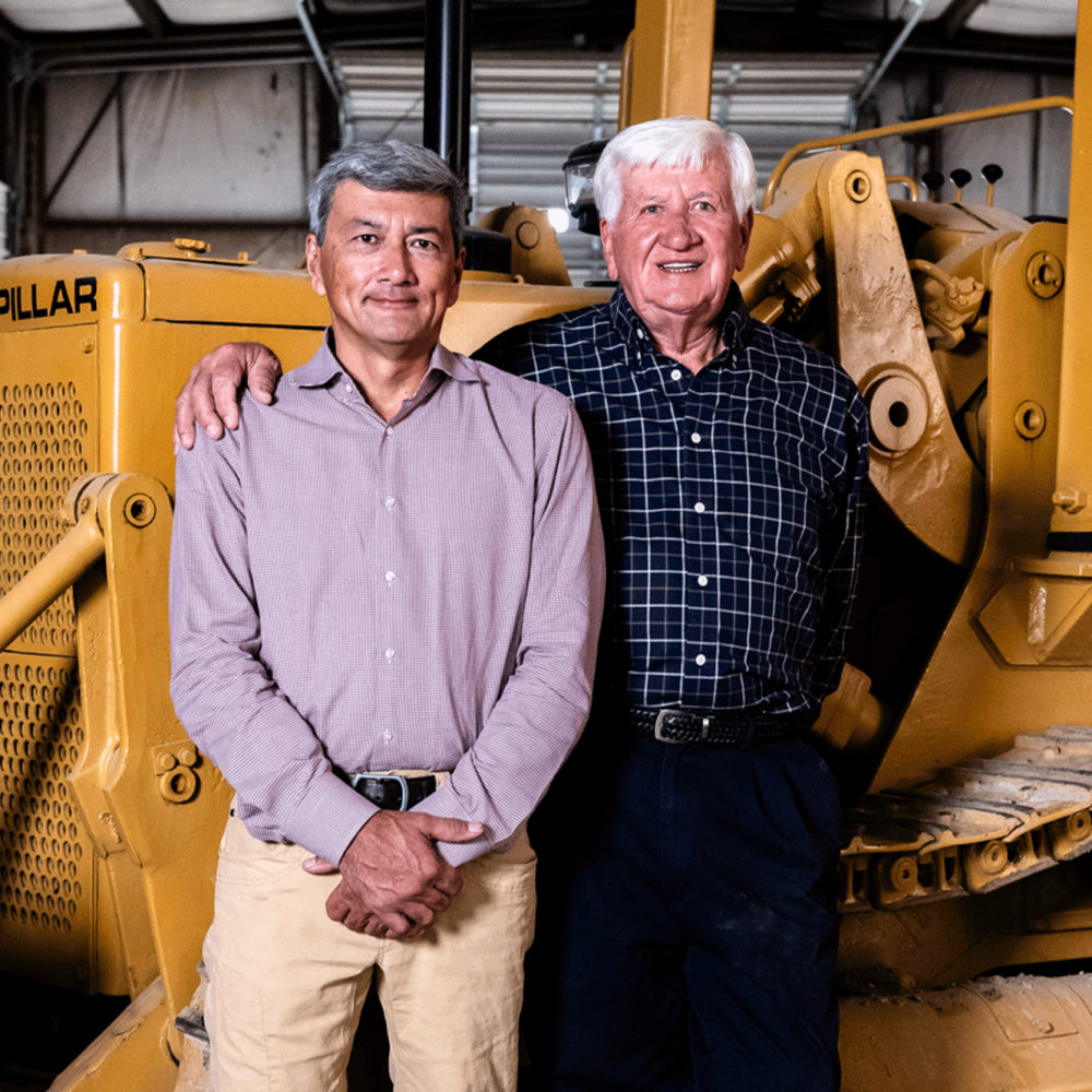 Two men stand by a yellow Caterpillar bulldozer, one arm around other.