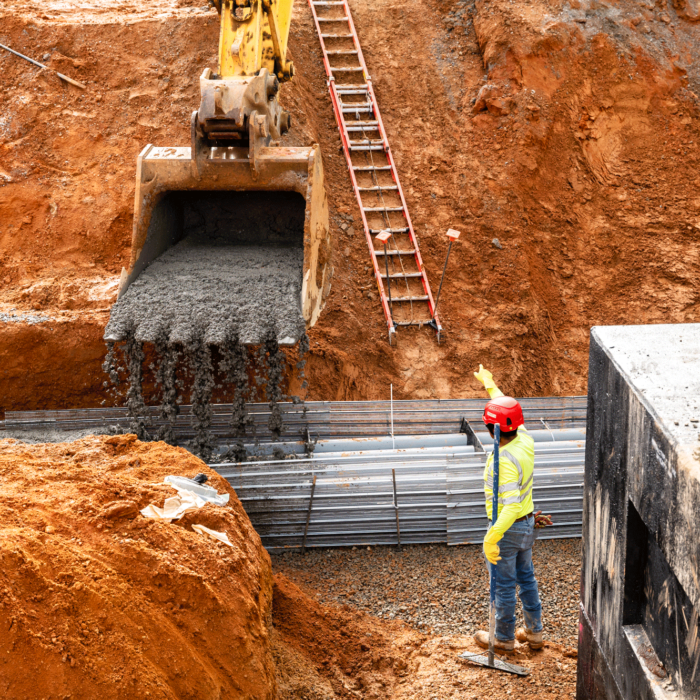 Worker guides excavator pouring gravel; steel beams, ladder behind.