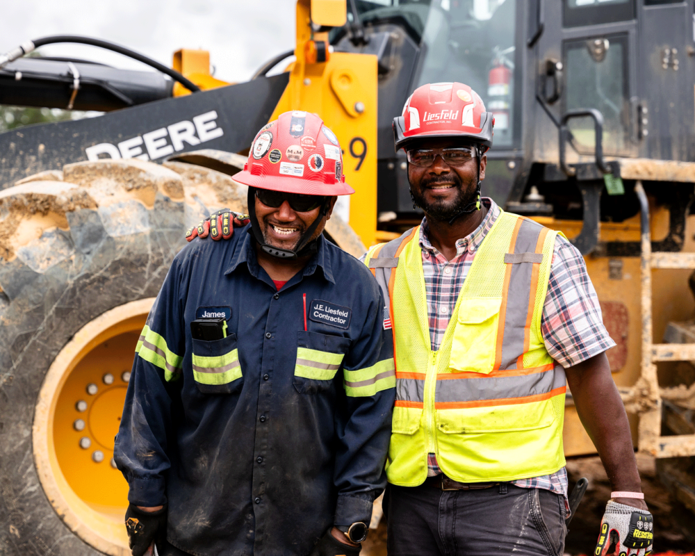 Two construction workers in safety gear smile by machinery.