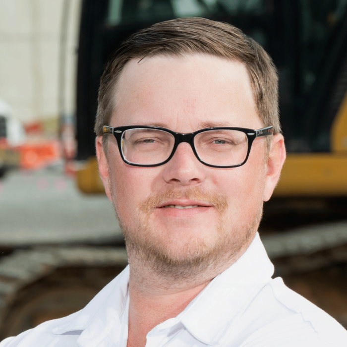 Man with glasses and white shirt stands by construction equipment.