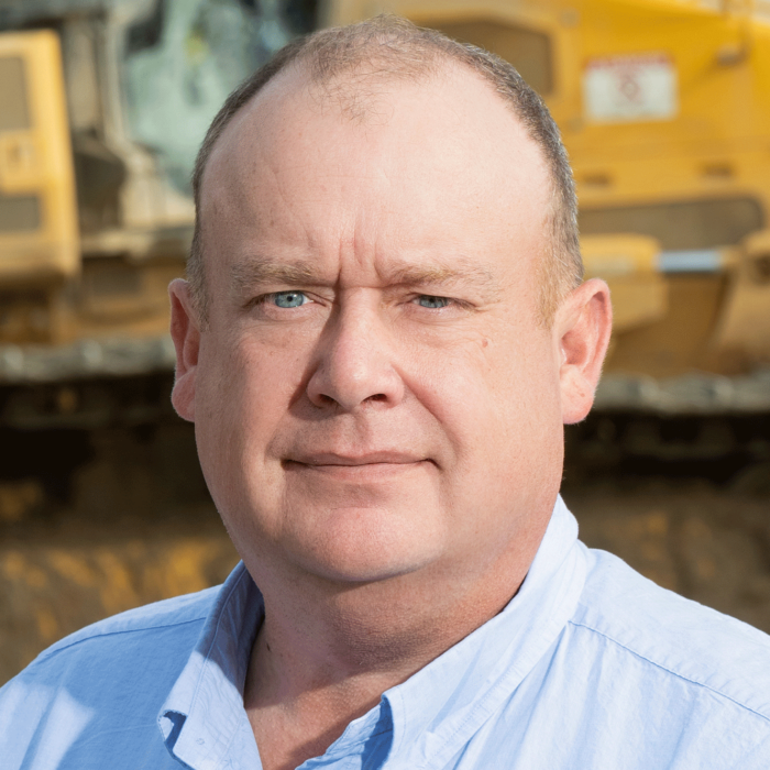 Man in blue shirt stands by construction equipment, facing camera.