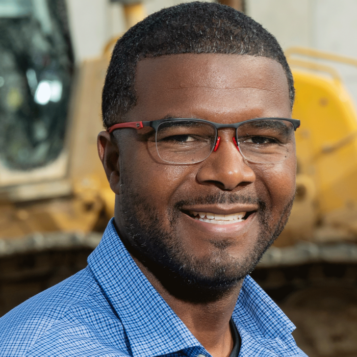 Man in glasses, blue check shirt, smiles by construction equipment.