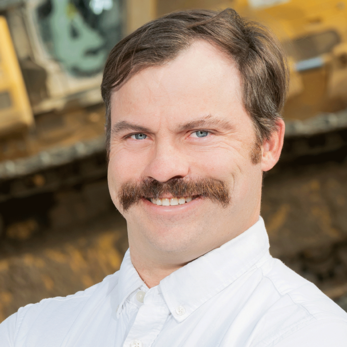 Smiling man with mustache stands before construction equipment.