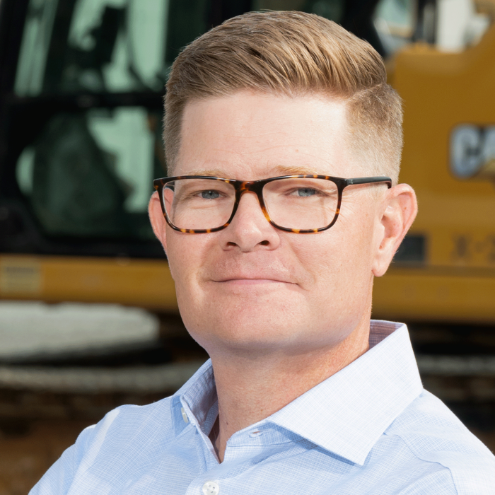 Man with glasses stands by a yellow construction vehicle.