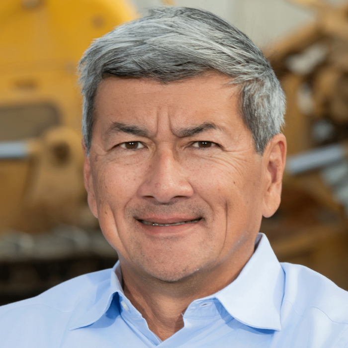 Smiling man in blue shirt, construction equipment blurred behind.