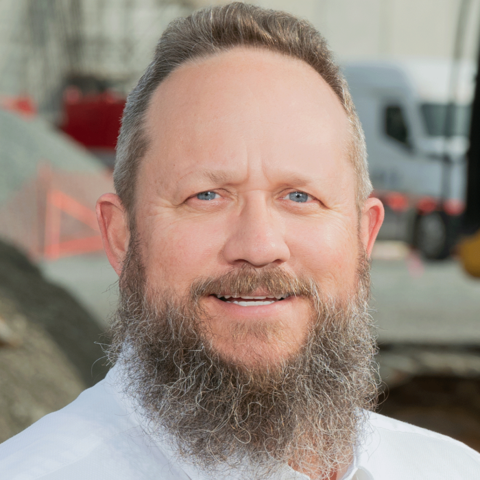 Bearded man in white shirt stands at outdoor construction site.