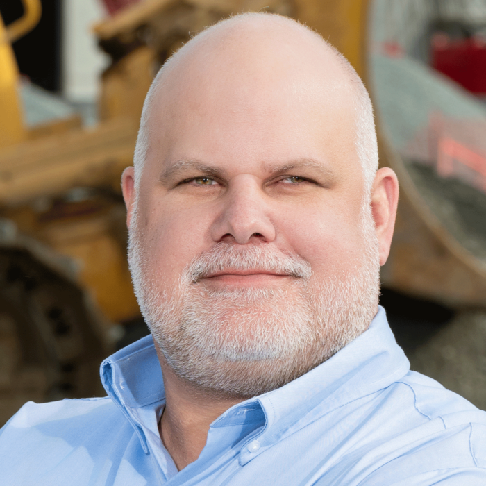 Bearded man in blue shirt stands by construction equipment.