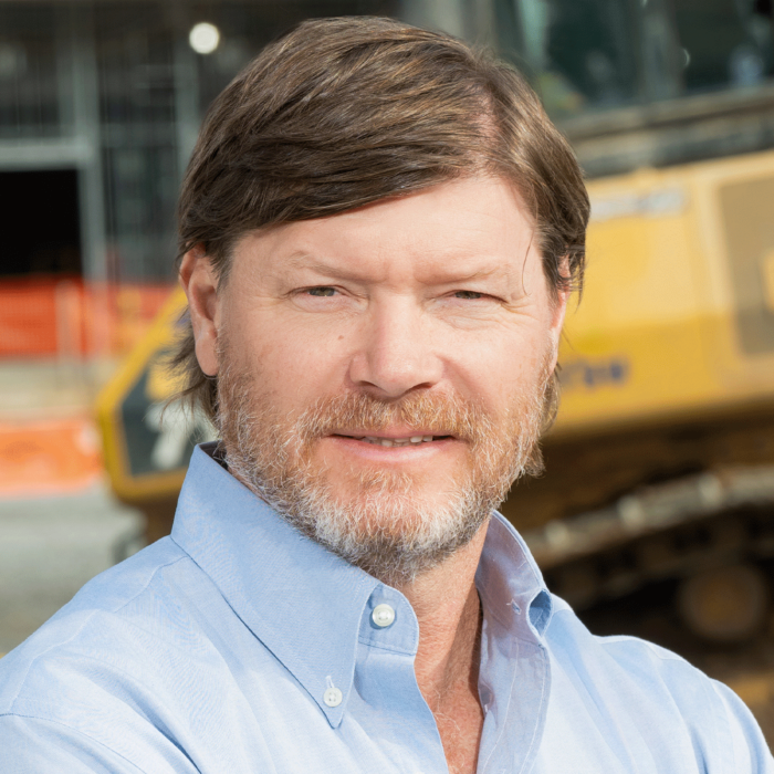 Bearded man in blue shirt stands by construction equipment outdoors.