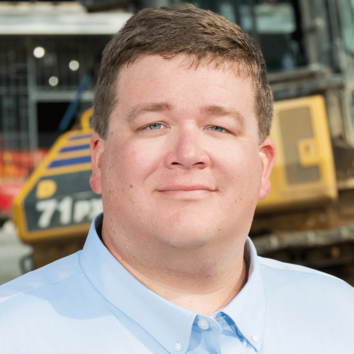 Man in light blue shirt stands before construction site equipment.
