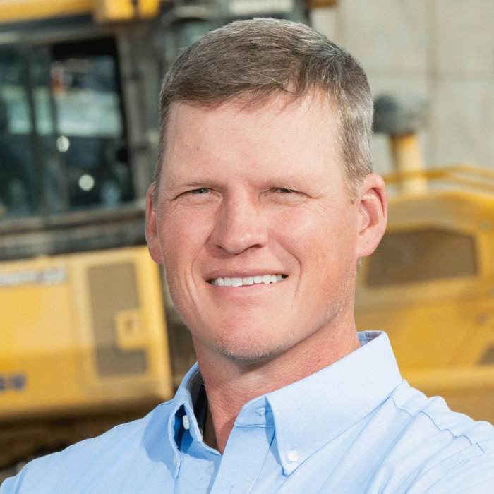 Man in light blue shirt stands before yellow construction equipment.