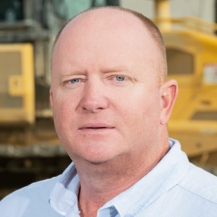Man in light blue shirt stands before yellow construction equipment.