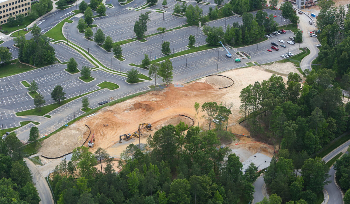 Aerial view: construction site, parking lot, buildings, trees nearby.