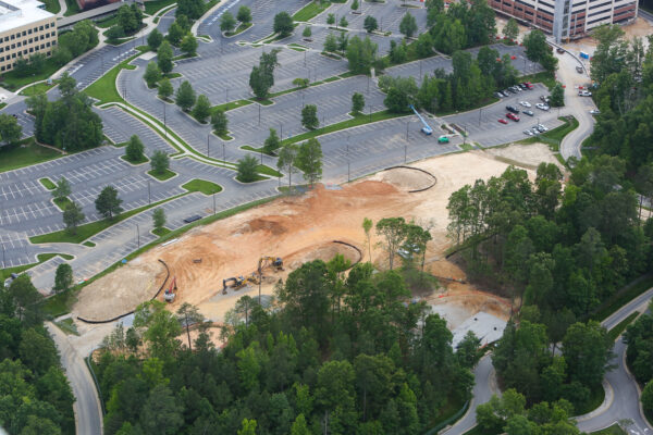 Aerial view: construction site, parking lot, buildings, trees nearby.