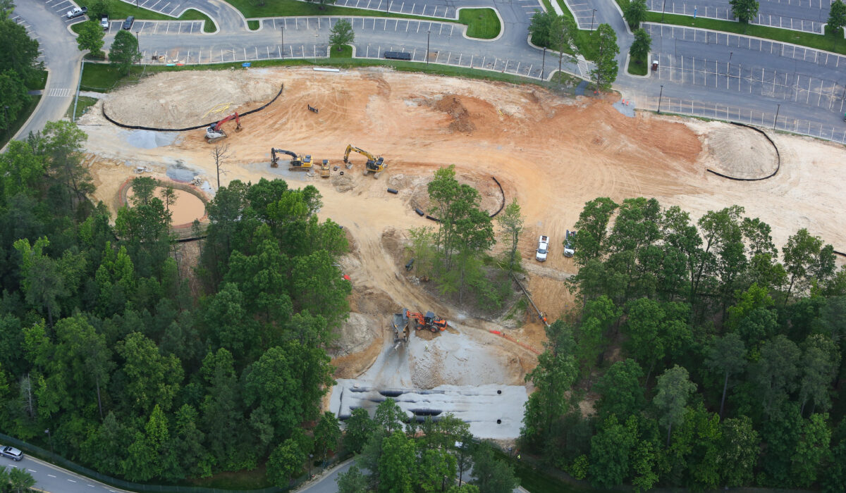 Aerial view: construction site, excavators, vehicles, trees, empty lot.