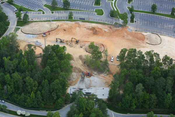 Aerial view: construction site, excavators, vehicles, trees, empty lot.