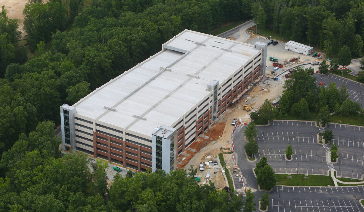 Multi-level parking garage under construction, trees and vehicles nearby.