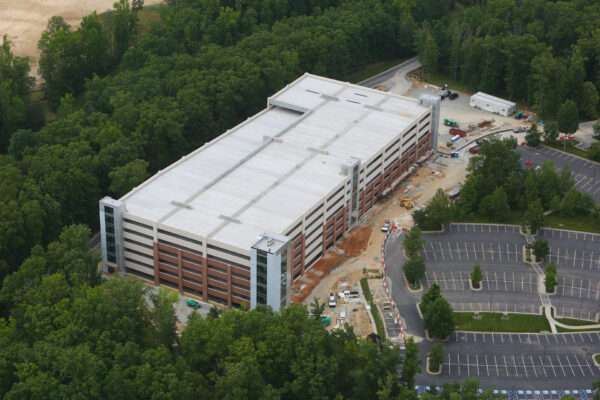 Multi-level parking garage under construction, trees and vehicles nearby.