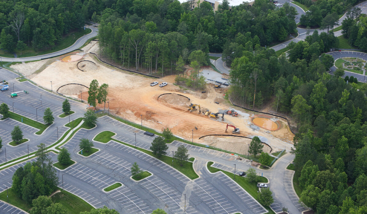 Aerial view: construction site, excavators, vehicles, parking lot, trees.