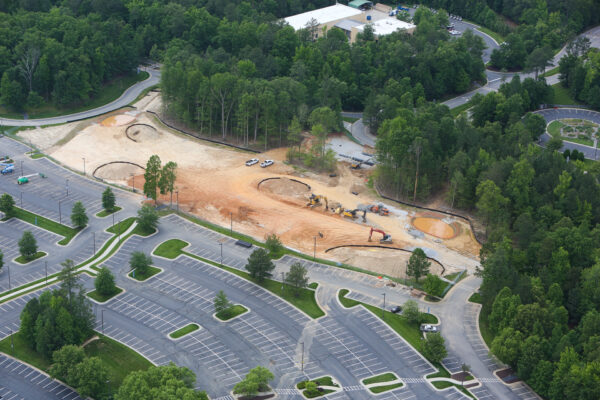 Aerial view: construction site, excavators, vehicles, parking lot, trees.