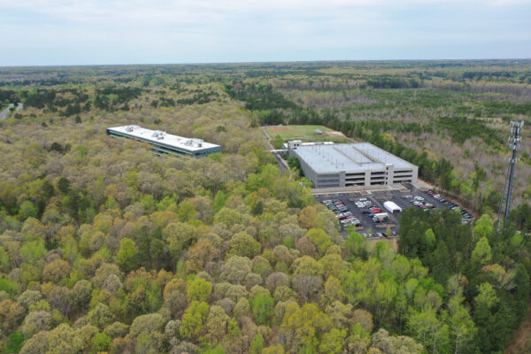 Aerial view: two office buildings, forest, parking lot, cars.