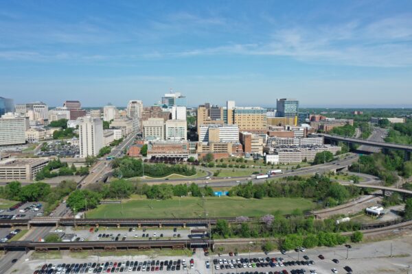 Aerial cityscape: tall buildings, highways, green spaces, blue sky.