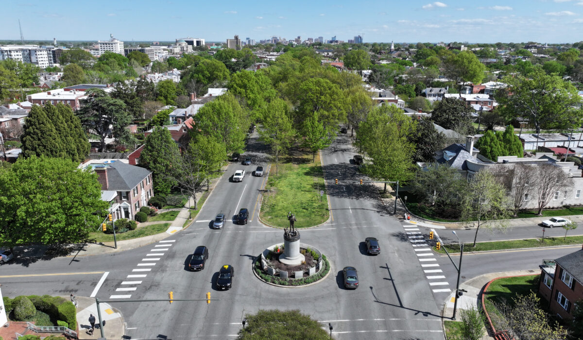 Aerial view: roundabout with statue, trees, homes, distant skyline.
