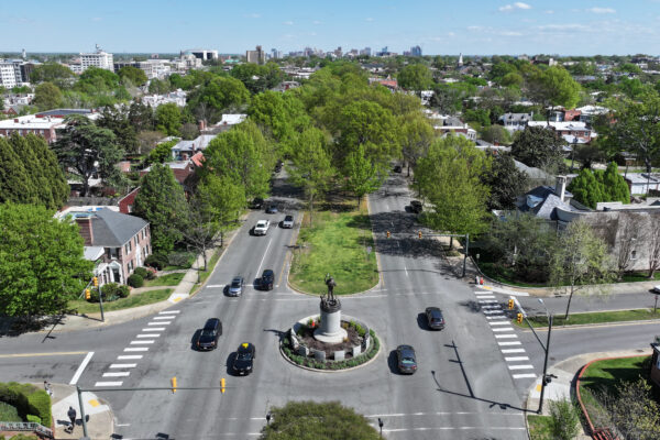 Aerial view: roundabout with statue, trees, homes, distant skyline.