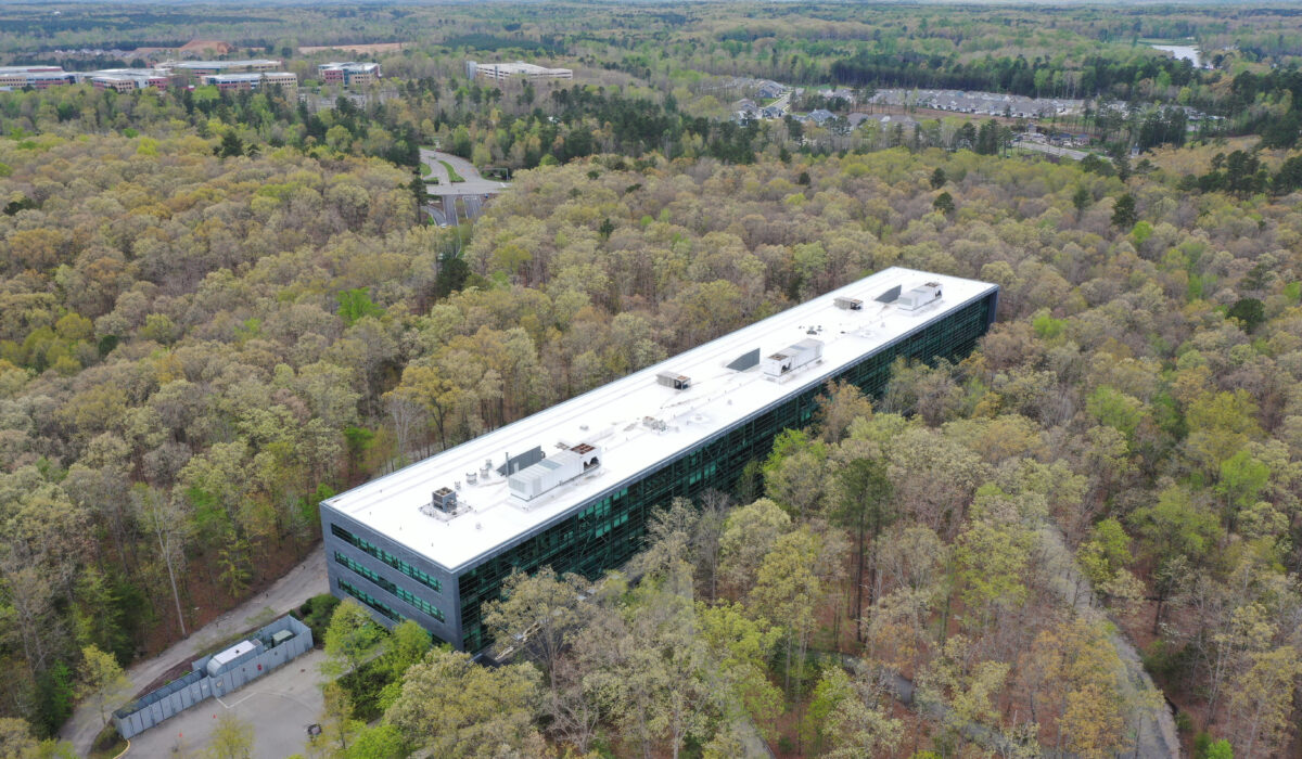 Aerial view: white-roofed building in forest, buildings behind.