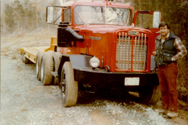 Man stands by red Autocar semi on gravel path, trees nearby.