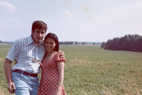 A couple stands in a field; man’s arm around woman.