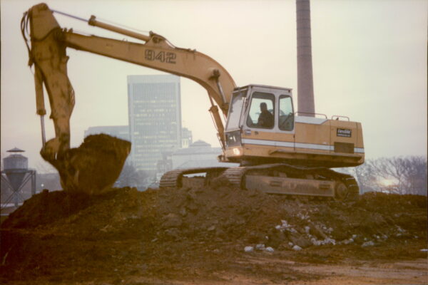 Yellow excavator moves dirt; buildings and smokestack behind.