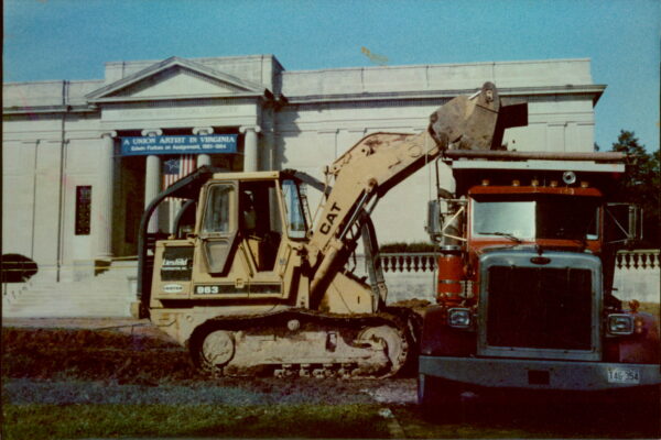 Yellow excavator and red dump truck parked before columned building.