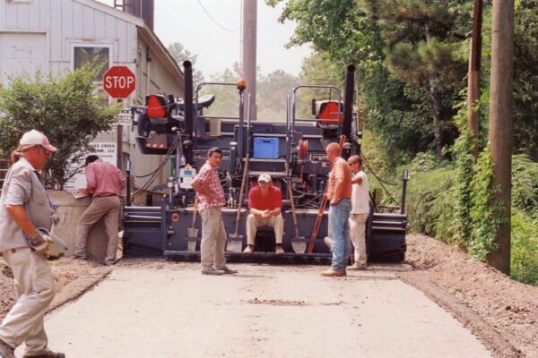 Construction workers work near paving machine; stop sign in background.