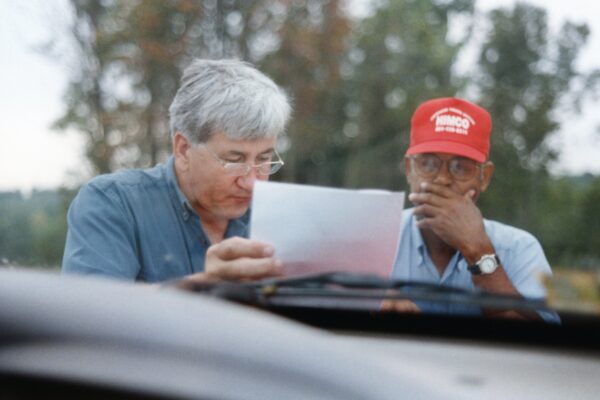 Two men review a document outdoors; trees in background.