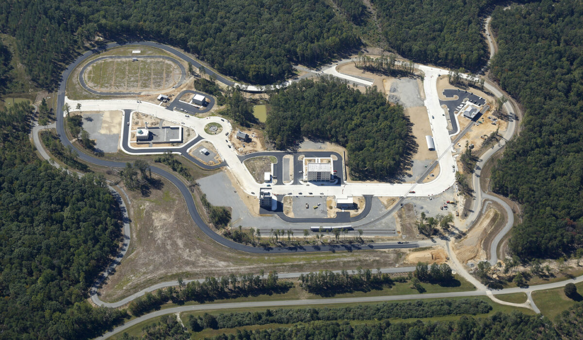 Aerial view of fenced facility with buildings and roads in forest.