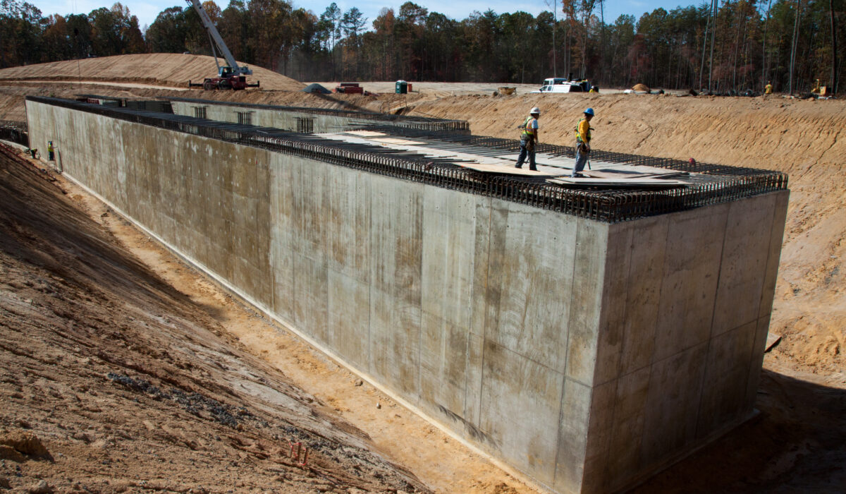 Two workers stand atop a large concrete structure, forest behind.