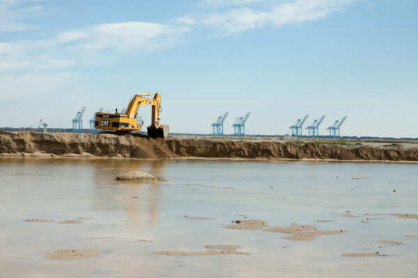 Yellow excavator digs near water; cranes in the background.