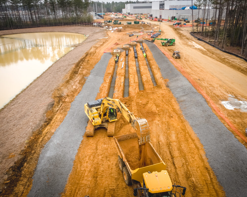 Machinery digs parallel trenches near a pond, trees, buildings.