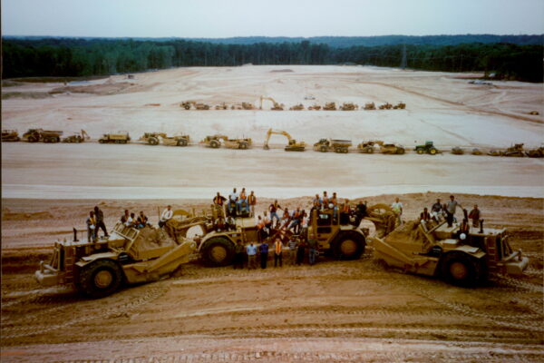 Crowd with earthmovers at construction site, forest in background.