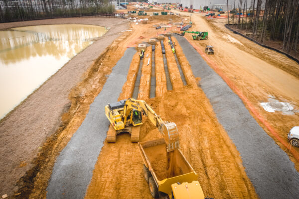 Heavy machinery digging trenches; trees, pond, and building nearby.
