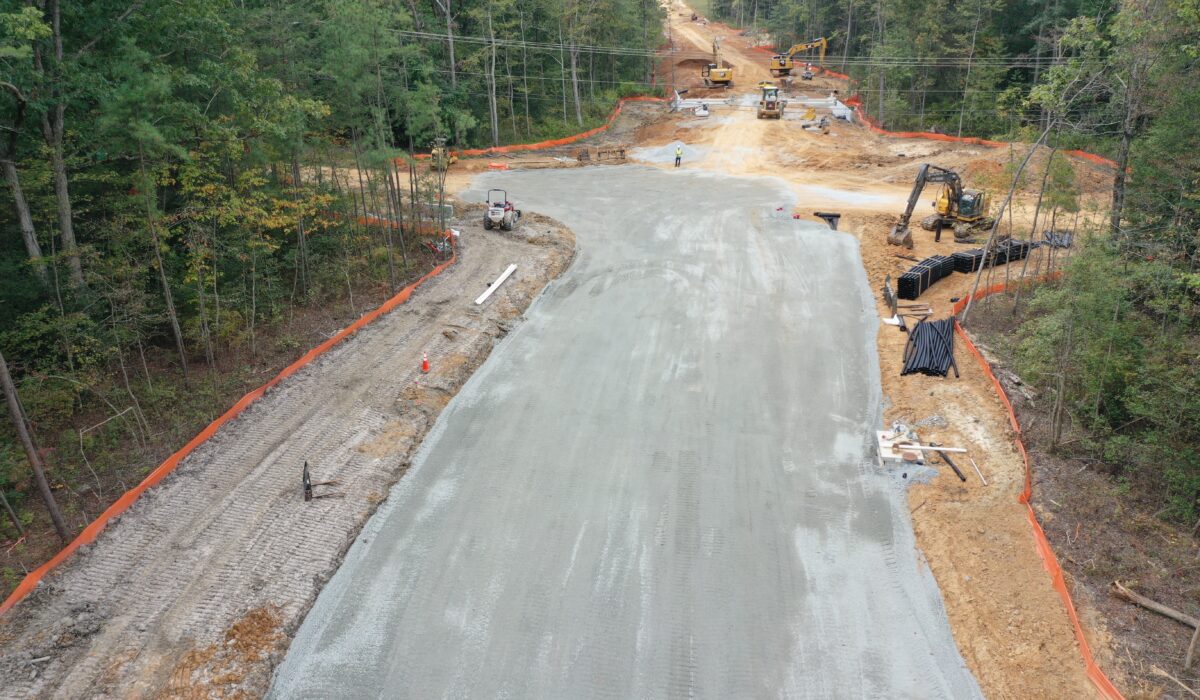 Workers and machines paving road through woods, orange fencing borders.