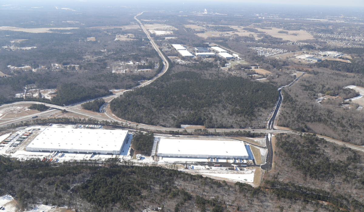 Aerial view: two white-roof warehouses, trees, roads, snow patches.