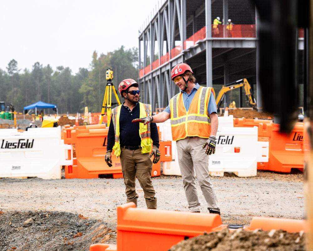 Two workers in safety gear talk at a construction site.